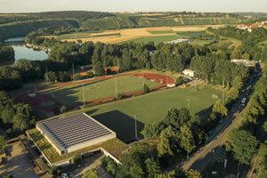 Gustav-Siegle-Stadion Luftaufnahme eines Sportgeländes mit Fußball- und Tennisplätzen, umgeben von Bäumen und einem Fluss im Hintergrund.