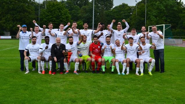 Mannschaftsfoto einer Fußballmannschaft in weißen Shirts mit "Aufsteiger Bezirksliga 24", viele feiern mit einer Trophäe.