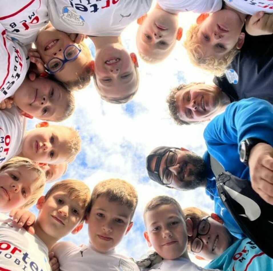 Gruppe von Kindern und Erwachsenen, die nach oben schauen und lächeln, umrahmt von blauem Himmel.