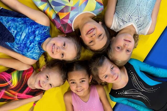 Sechs lächelnde Kinder in bunten Turnanzügen liegen auf einer gelben Turnmatte und blicken in die Mitte.