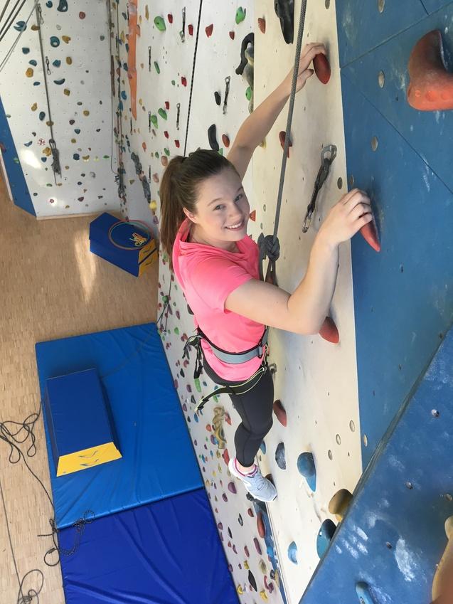 Junge Frau klettert an einer bunten Kletterwand mit Sicherungsgurt und lächelt, blauer Untergrund mit Matten.