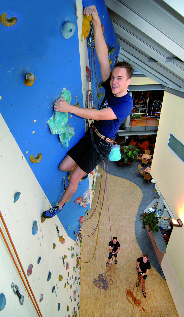 Junger Mann klettert an einer blauen Kletterwand, während zwei Personen unten mit Seilen sichern. Indoor-Kletterhalle.
