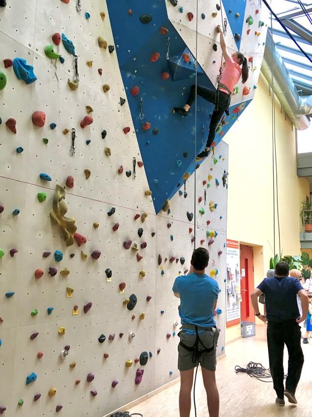Eine Person klettert an einer kunterbunten Kletterwand, während eine weitere Person den Seilzug hält.