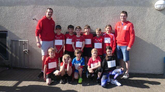 Gruppe von Kindern in roten T-Shirts mit einem Trainer, posierend vor einer Wand, bei einem Sportereignis.