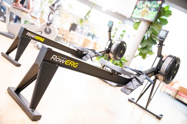 Nahaufnahme von zwei Ruderergometern in einem Fitnessstudio mit einem Bildschirm im Hintergrund.