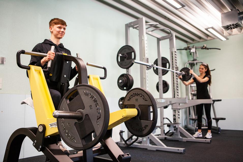 Junger Mann trainiert an einer Kraftmaschine, während eine Frau mit einer Langhantel im Hintergrund trainiert.