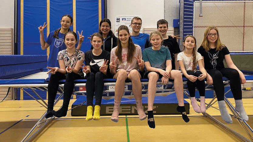 Gruppe von zehn Kindern und Jugendlichen sitzt auf einem Trampolin, lächelnd und mit V-Zeichen.