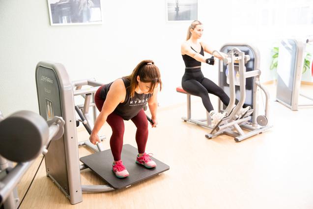 Zwei Frauen trainieren in einem Fitnessstudio an Geräten; eine macht Kniebeugen, die andere sitzt und zieht.