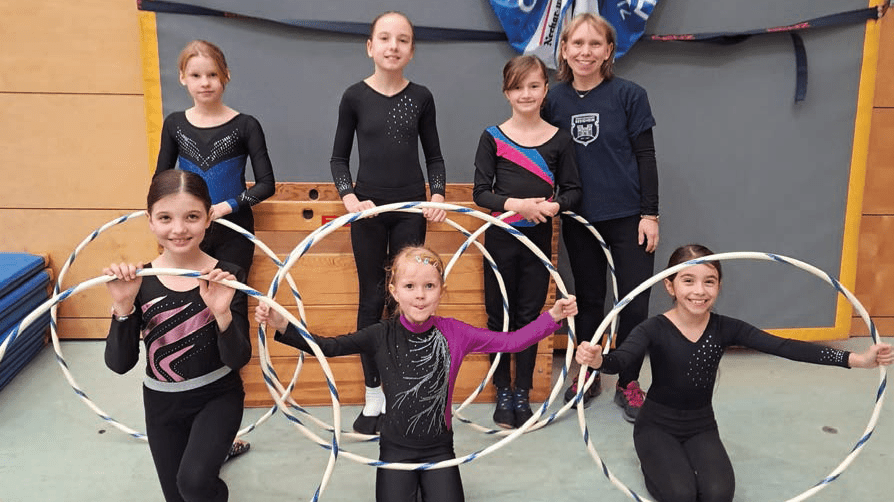 Sechs Mädchen in Sportoutfits halten Hula-Hoops, umringt von Trainingsgeräten in einer Turnhalle.