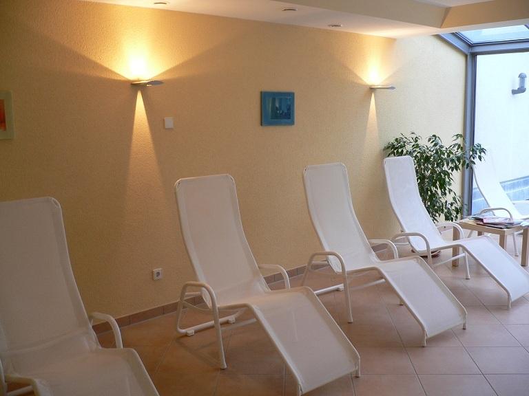 Ruhebereich mit fünf weißen Liegen, sanft beleuchtet, einer Pflanze und Fensterelementen.