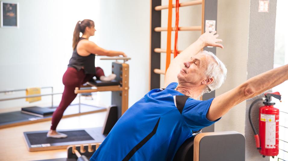 Senior in blauer Sportbekleidung dehnt sich auf einem Fitnessgerät, während eine Frau im Hintergrund trainiert.