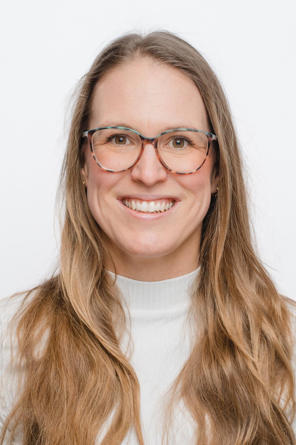 Porträt einer lächelnden Frau mit langen, lockigen Haaren und einer weißen Bluse vor einem weißen Hintergrund.