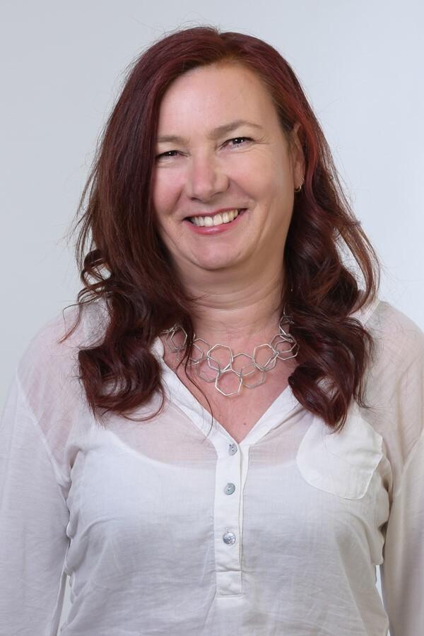 Frau mit langen, lockigen roten Haaren, trägt eine weiße Bluse und eine silberne Kette, lächelt in die Kamera.