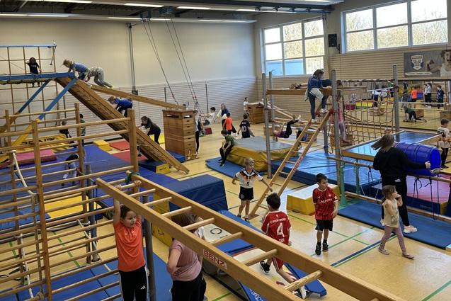 verschiedene Geräte in der Sporthalle sind aufgebaut. Kinder toben darauf