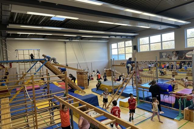 verschiedene Geräte in der Sporthalle sind aufgebaut. Kinder toben darauf