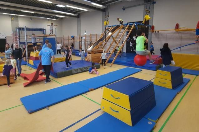 Kinder turnen in einer Sporthalle an verschiedenen Geräten wie Matten, einem Klettergerüst und einer Rutsche.