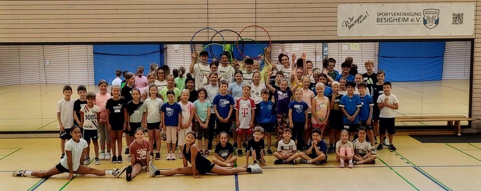 Gruppenfoto von Kindern in Sportkleidung, einige mit Namensschildern, in einer Sporthalle mit Olympiaringen im Hintergrund.