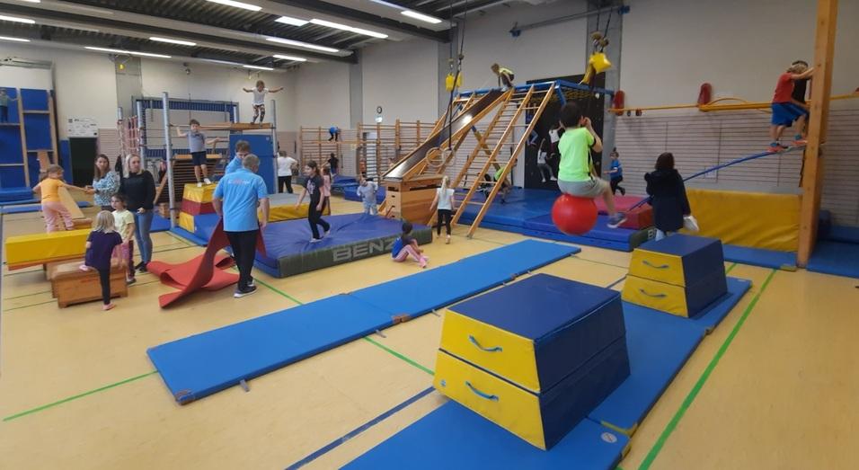 Kinder turnen in einer Sporthalle an verschiedenen Geräten wie Matten, einem Klettergerüst und einer Rutsche.