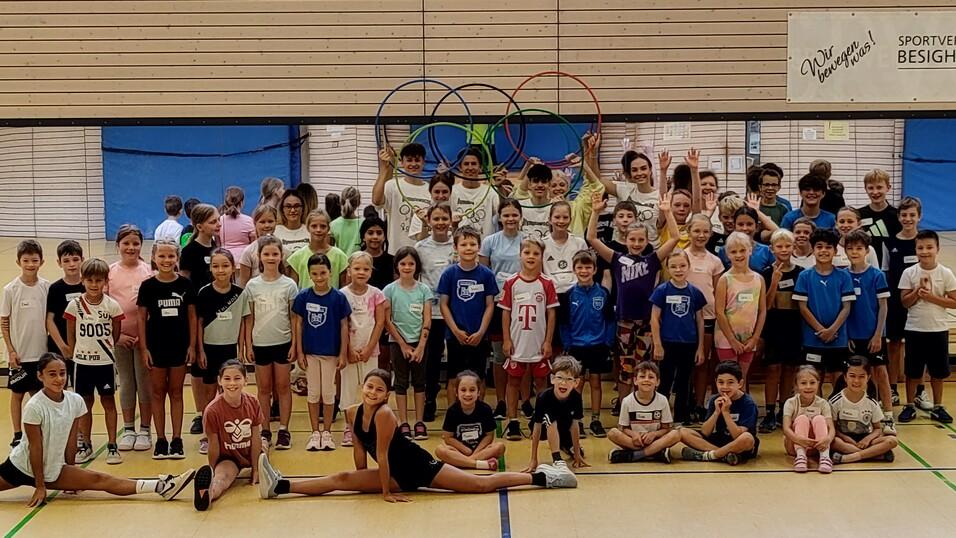 Gruppenfoto von Kindern in Sportkleidung, einige mit Namensschildern, in einer Sporthalle mit Olympiaringen im Hintergrund.