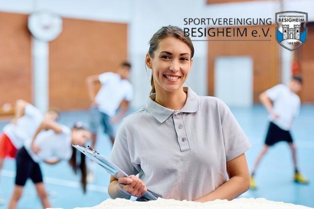 Lächelnde Frau mit Clipboard steht im Vordergrund, während im Hintergrund eine Gruppe in einer Sporthalle Übungen macht.