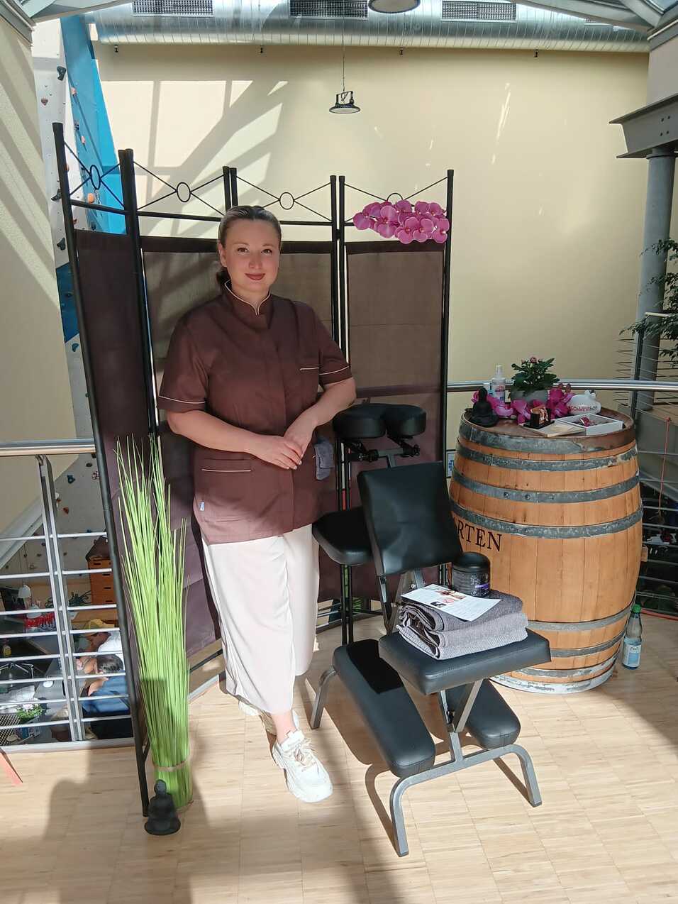 Frau in beiger Kleidung steht neben einem Sichtschutz und einer Massageliege mit Handtüchern in einem hellen Raum.