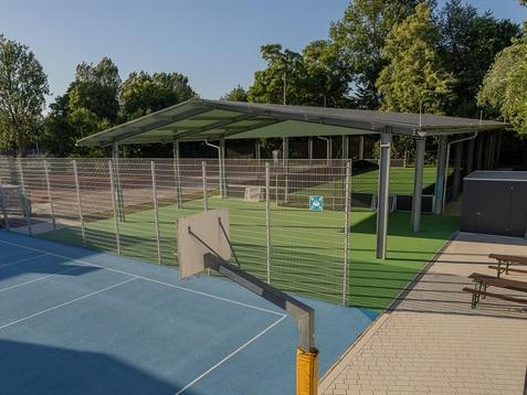 Sportanlage mit blauer Spielfläche, Basketballkorb und überdachtem Bereich im Hintergrund, umgeben von Bäumen.
