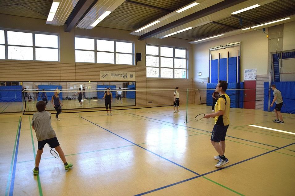 Sechs Personen spielen Badminton in einer Sporthalle mit Fensterfront und zwei Netzen.