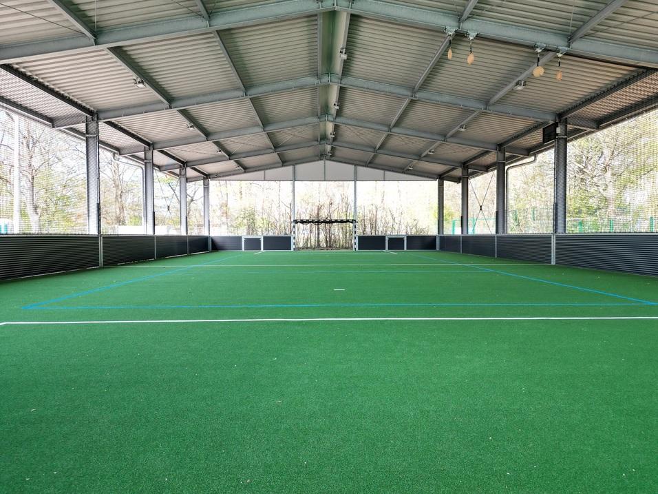 Überdachte Sporthalle mit grünem Kunstrasen und Tor auf einer Seite, umgeben von Netzen und Fenstern.