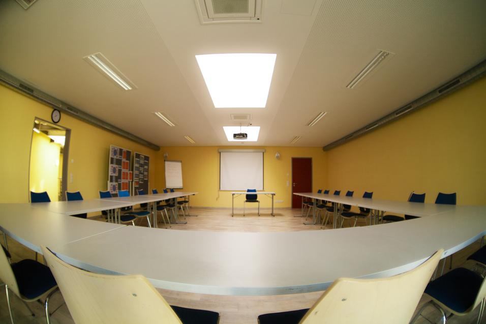 Konferenzraum mit runder Tischanordnung, blauen Stühlen, Projektor und gelben Wänden. Beamerleinwand in der Mitte.