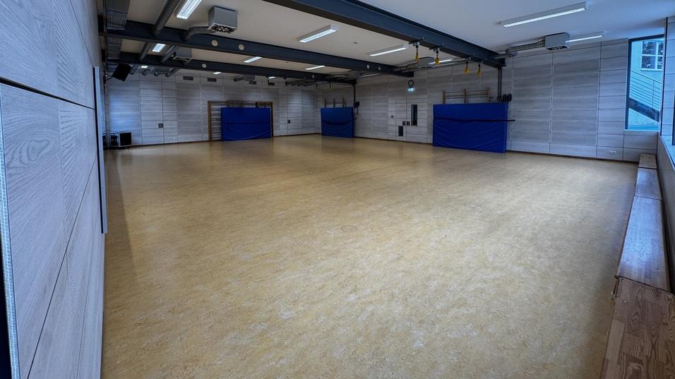 Leere Sporthalle mit braunem Boden, blauen Sichtschutzwänden und Holzbank entlang der Wand. Tageslicht durch Fenster.