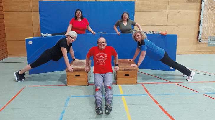 Fünf Personen in Sportkleidung machen verschiedene Übungen an Holzboxen in einer Sporthalle.