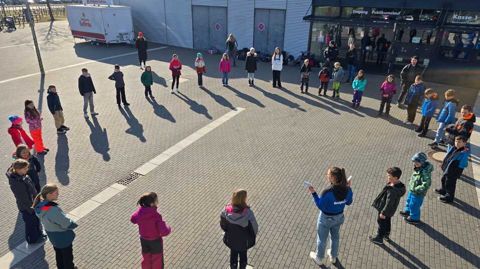 Gruppe von Kindern steht im Kreis, während eine Anleiterin in blauer Jacke ihnen Anweisungen gibt.
