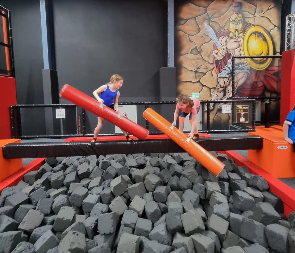 Zwei Kinder kämpfen mit Schaumstoffstangen auf einer Wippe über einem großen Schaumstoffblock-Bereich.