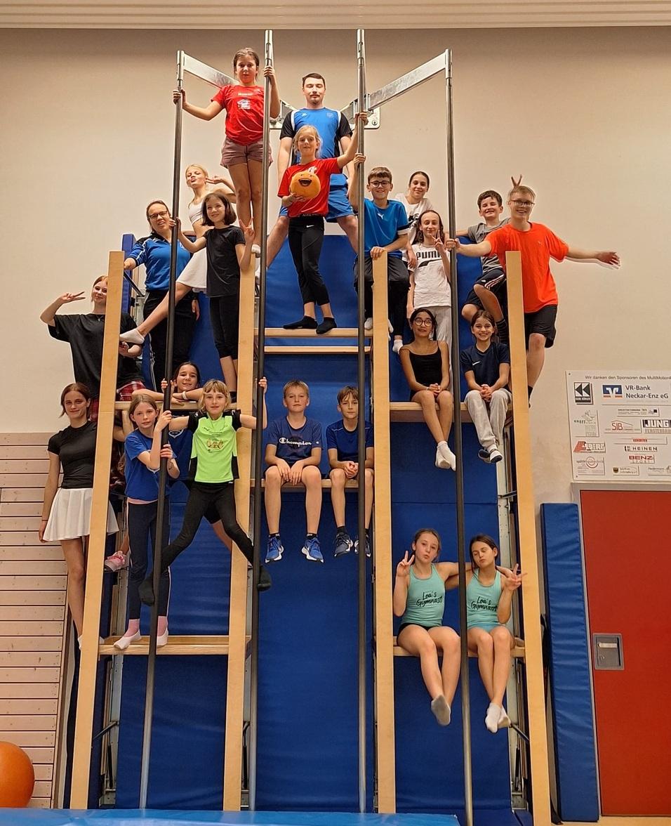 Gruppengymnastik mit Kindern und Jugendlichen auf einem Klettergerüst in einer Halle, manche posieren, andere sitzen.