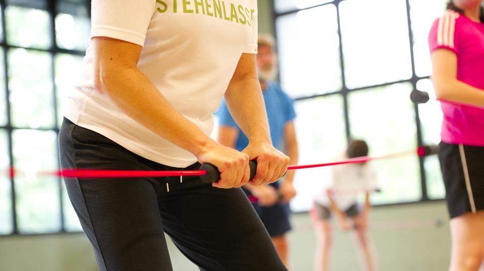 Nahaufnahme einer Person mit einem Widerstandsband im Fitnessstudio, umgeben von weiteren trainierenden Personen.