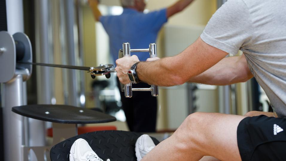 Mann trainiert im Fitnessstudio mit einem Kabelzuggerät, ein anderer Mensch im Hintergrund führt eine Übung aus.