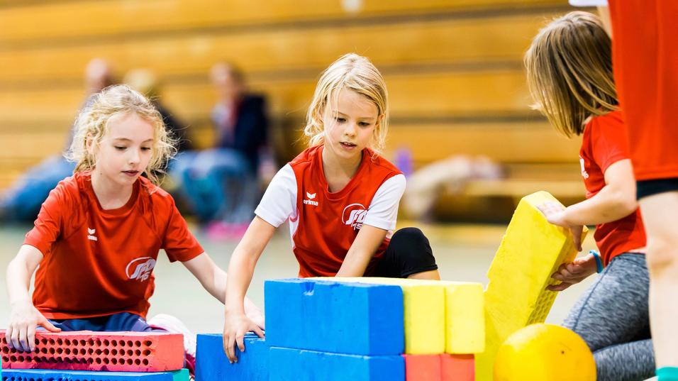 Drei Mädchen im roten Shirt bauen mit bunten Schaumstoffbausteinen in einer Turnhalle.