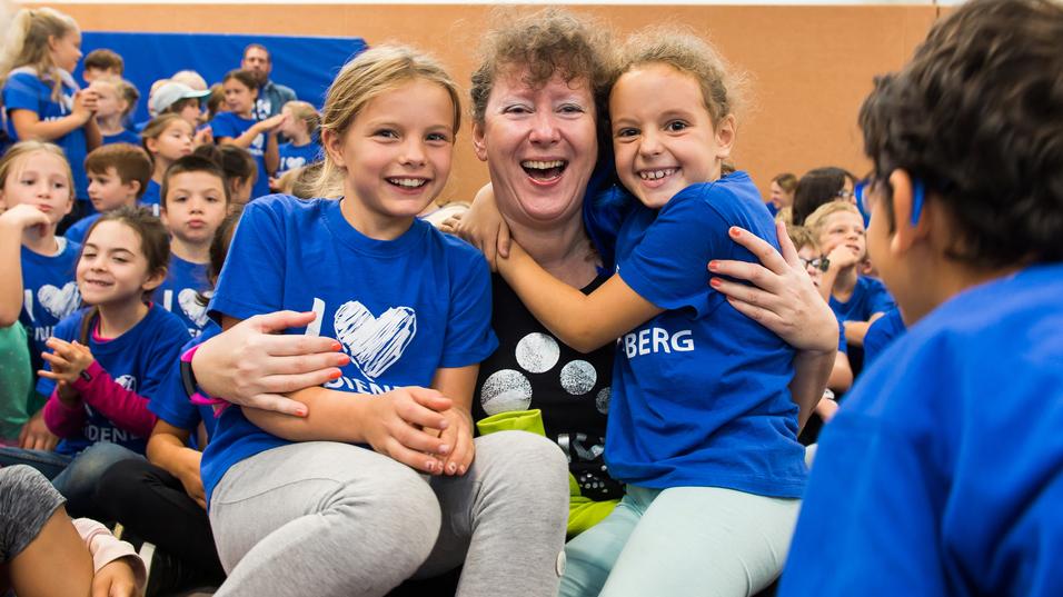 Lächelnde Kinder umarmen eine Frau in einem schwarzen Shirt, alle tragen blaue T-Shirts in einer Gruppenaktivität.