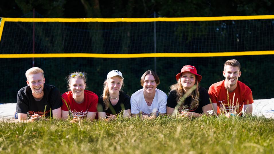 Sechs Personen liegen im Gras vor einem Beachvolleyballfeld mit gelben Netzen und Bäumen im Hintergrund.