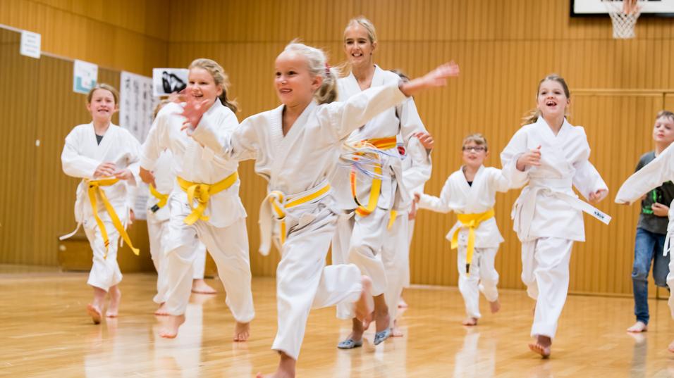 Gruppe von Kindern in weißen Karateanzügen mit gelben Gürteln, die fröhlich in einer Sporthalle herumrennen.