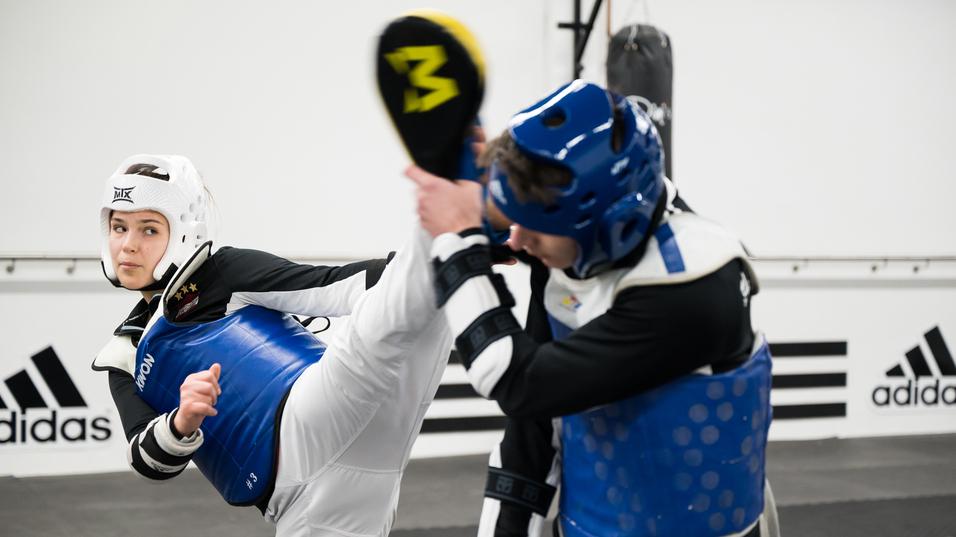 Zwei Kampfkämpfer im Taekwondo-Training; eine Person führt einen hohen Kick aus, beide tragen Schutzausrüstung.