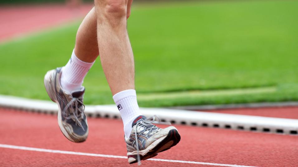 Läufer in grauen Sportschuhen hebt den Fuß auf einer roten Laufbahn über grünem Rasen.