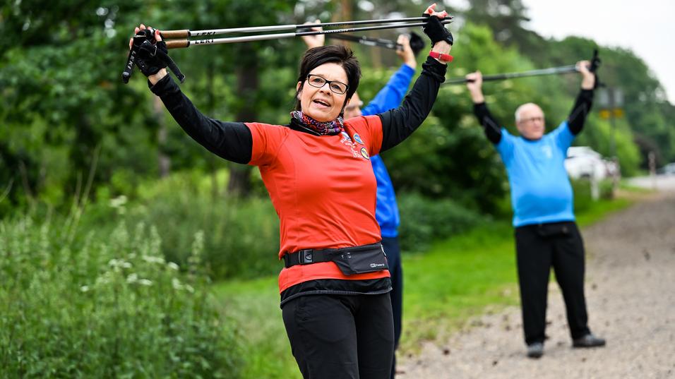 Frau in rotem Shirt hebt Wanderstöcke über den Kopf, während im Hintergrund zwei Personen in blauer und schwarzer Kleidung trainieren.