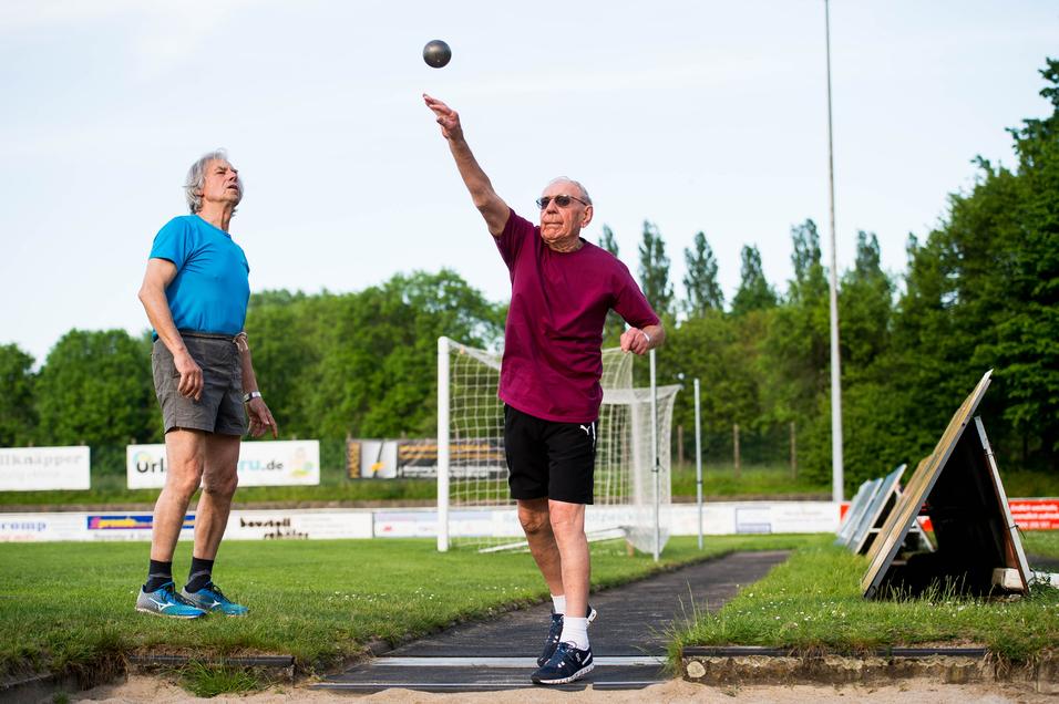 Zwei ältere Männer üben das Kugelstoßen auf einem Sportplatz, einer wirft die Kugel, der andere beobachtet.