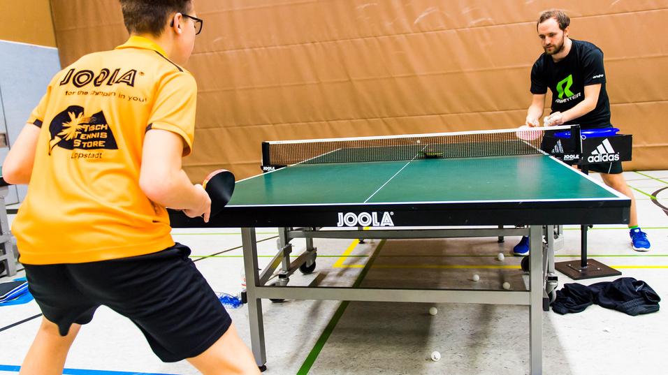 Zwei Tischtennisspieler trainieren an einem Tischtennistisch in einer Sporthalle, mehrere Bälle liegen auf dem Boden.