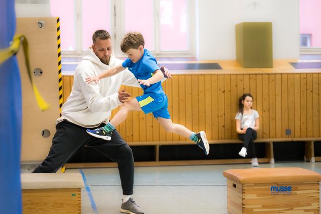 Ein junger Junge springt mit Hilfe eines Trainers über eine Trainingsbank in einer Turnhalle. Ein Mädchen beobachtet.