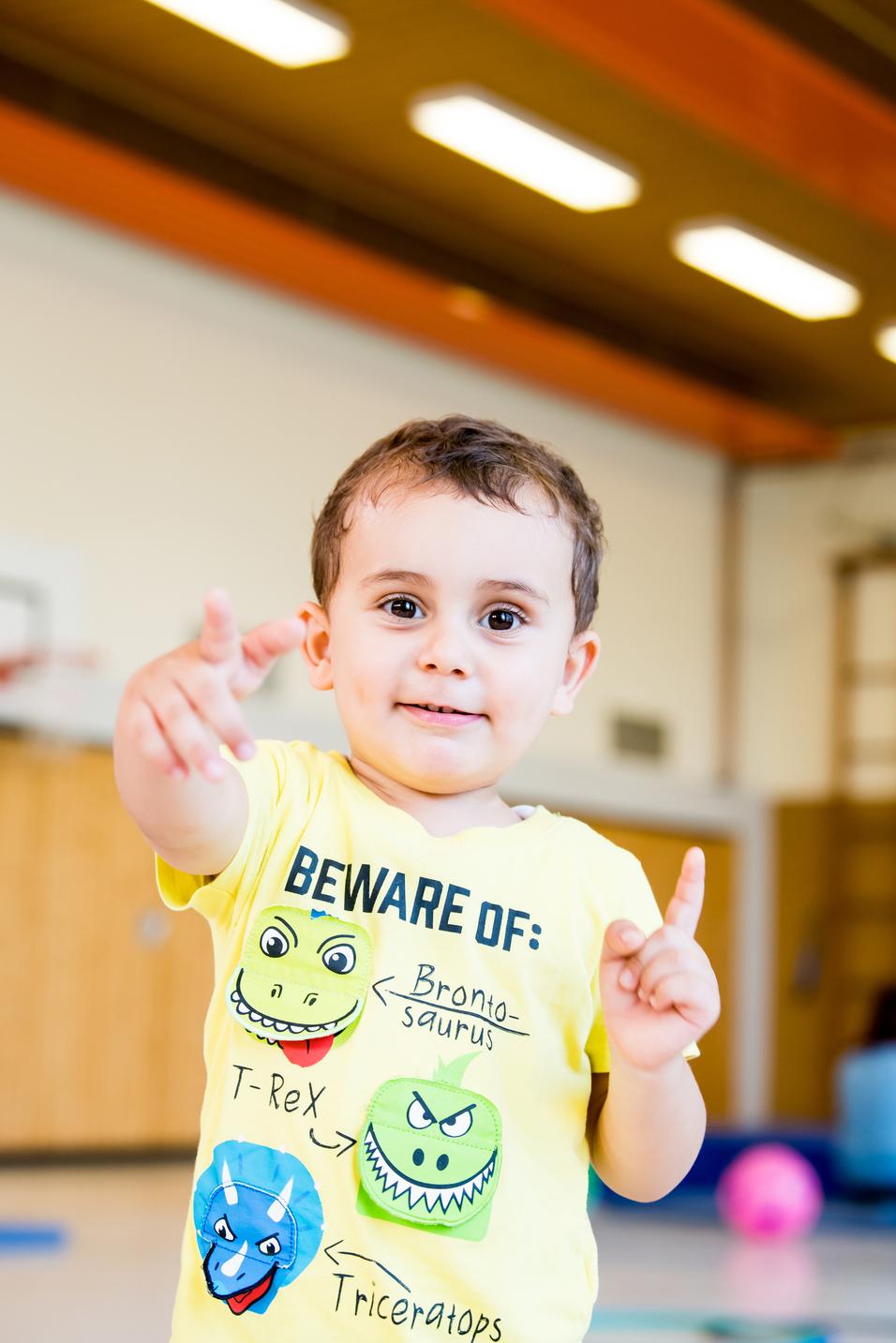 Lächelnder Junge in gelbem T-Shirt mit Dinosauriermotiven zeigt mit einem Finger auf einen Punkt im Raum.