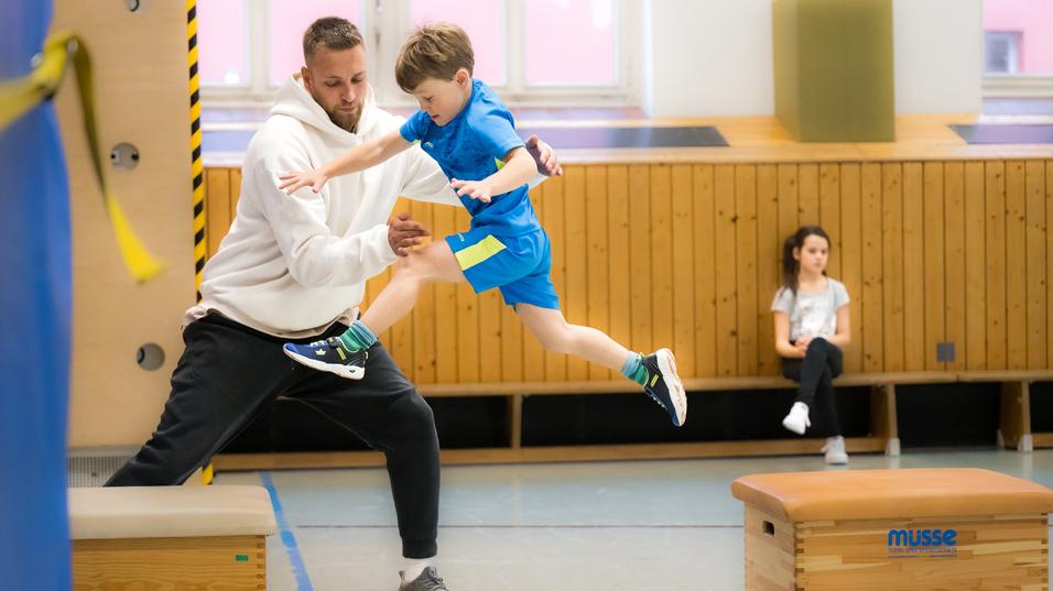 Ein junger Junge springt mit Hilfe eines Trainers über eine Trainingsbank in einer Turnhalle. Ein Mädchen beobachtet.