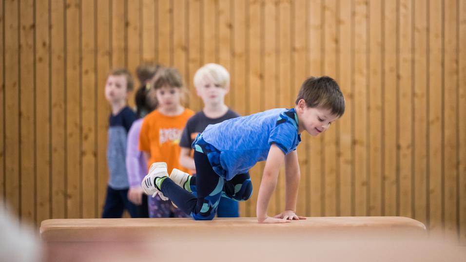 Ein Junge balanciert auf einem Blickkasten, während mehrere Kinder im Hintergrund zuschauen. Holzpaneele sind sichtbar.
