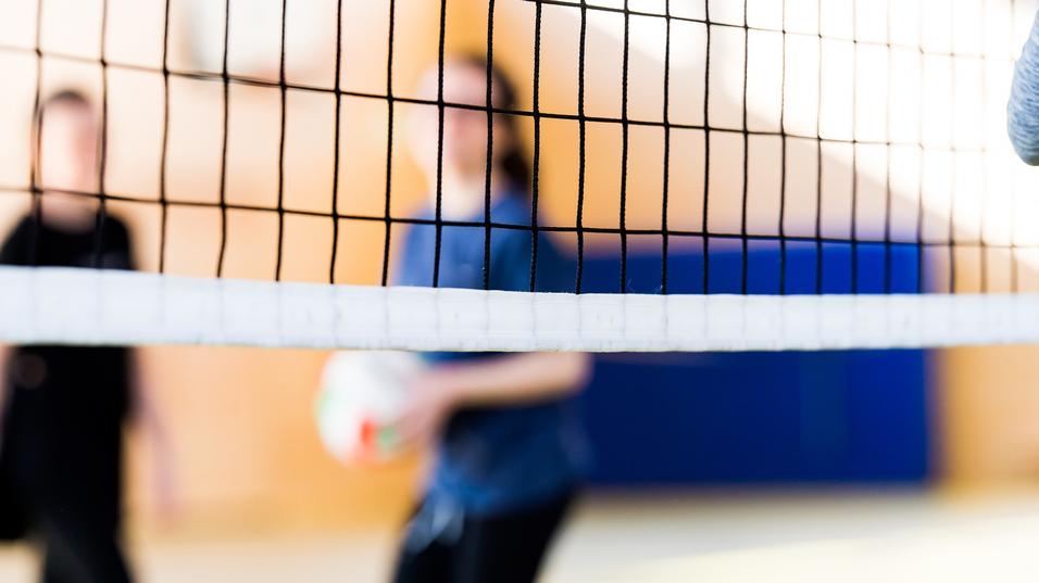 Unscharfe Silhouetten von zwei Personen hinter einem Volleyballnetz in einer Sporthalle. Eine Person hält einen Volleyball.
